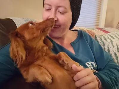 Carrie sits on a sofa as Nelly, a dog, looks into her eyes with love.